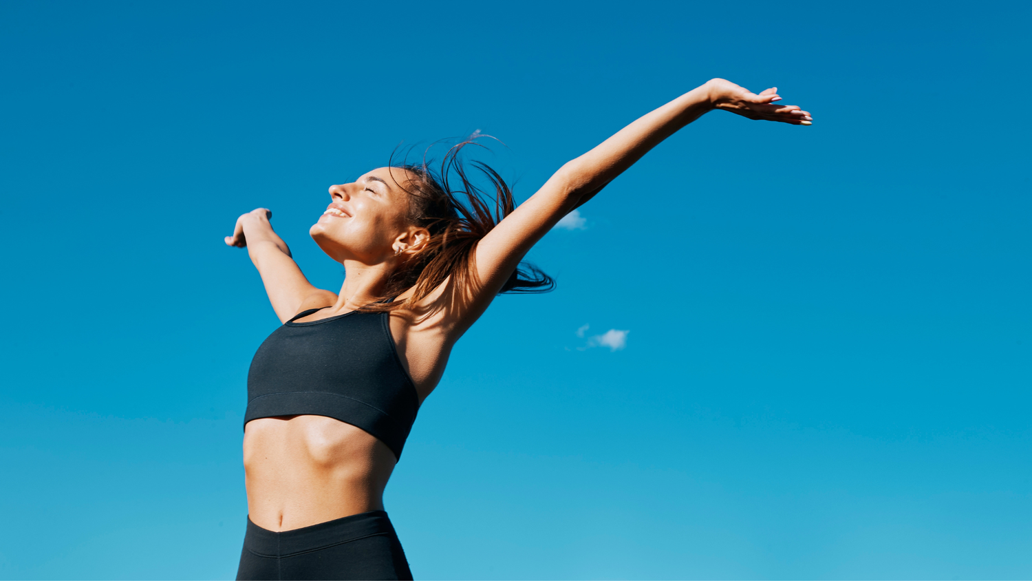 Clear blue sky with a female spreading her arms in the air smiling as the sun beams down on her face