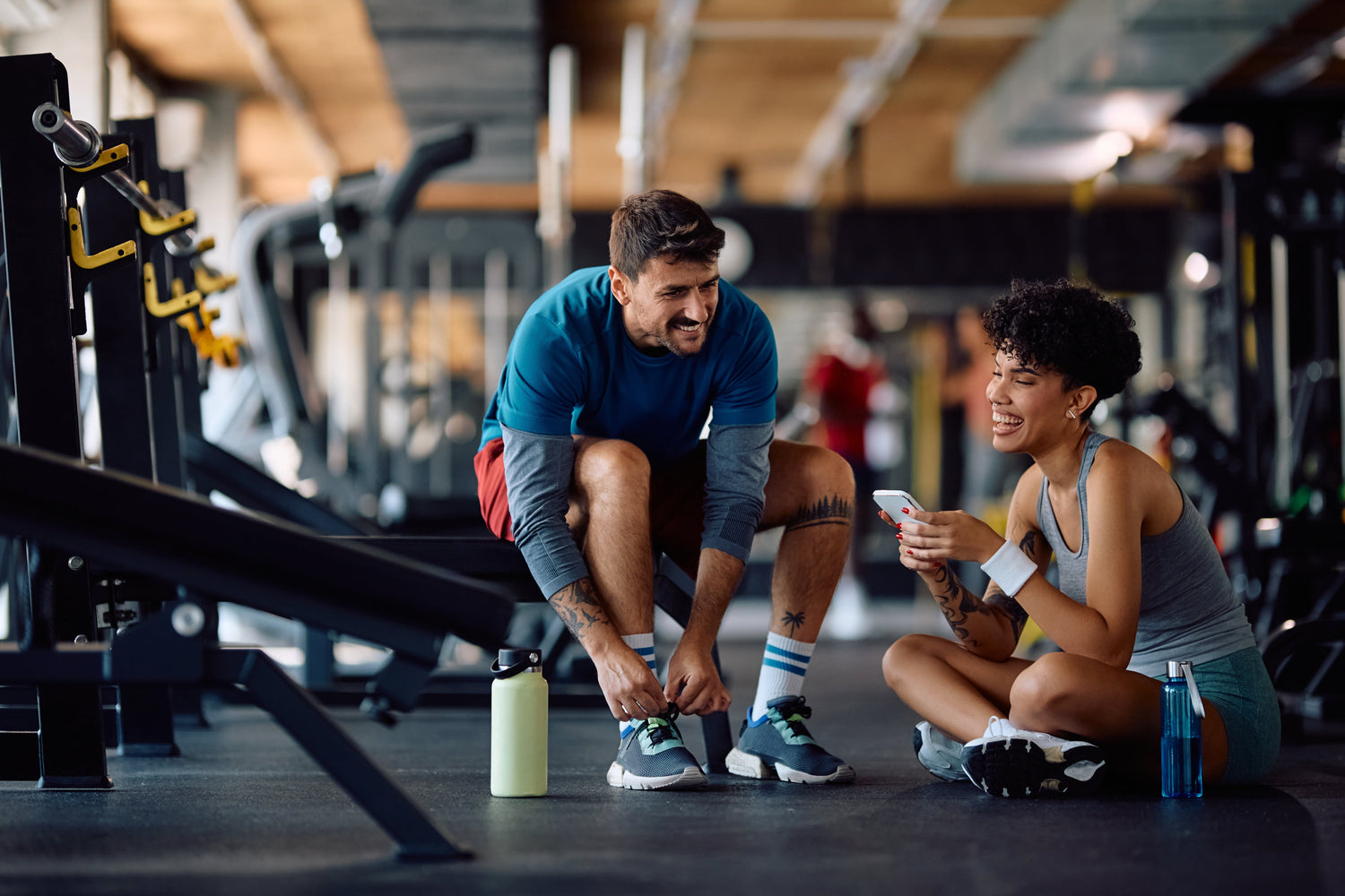 Gym setting 2 people, one sat on a bench the other sat on the floor laughing 
