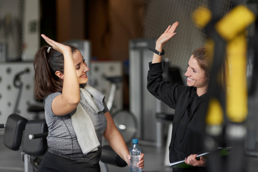 Two women in a gym setting, one with a towel on her shoulder and the other holding a water bottle, with gym equipment in the background.